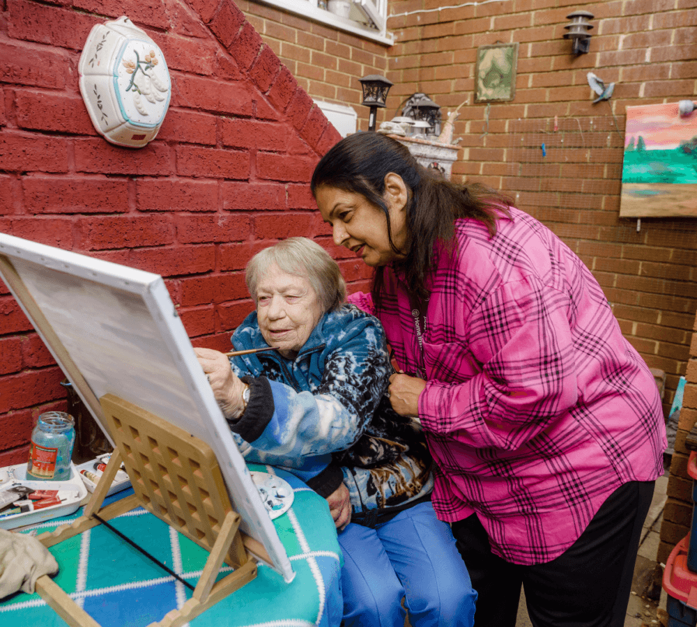An elderly woman painting on a canvas, assisted by another woman in a cozy, art-filled space. - Home Instead