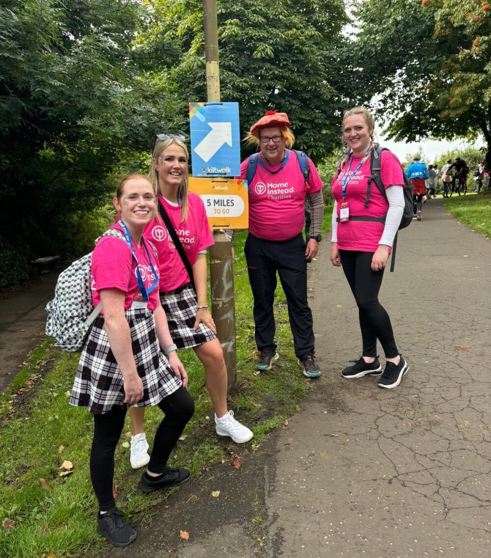 Four people in pink shirts smile by a signpost on a park path, surrounded by trees. - Home Instead