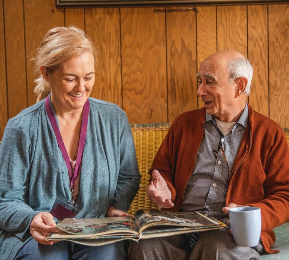 A carer wearing blue top smiling with a male senior chatting happily while looking at an album and having a cup of coffee sitting on a couch inside a house.