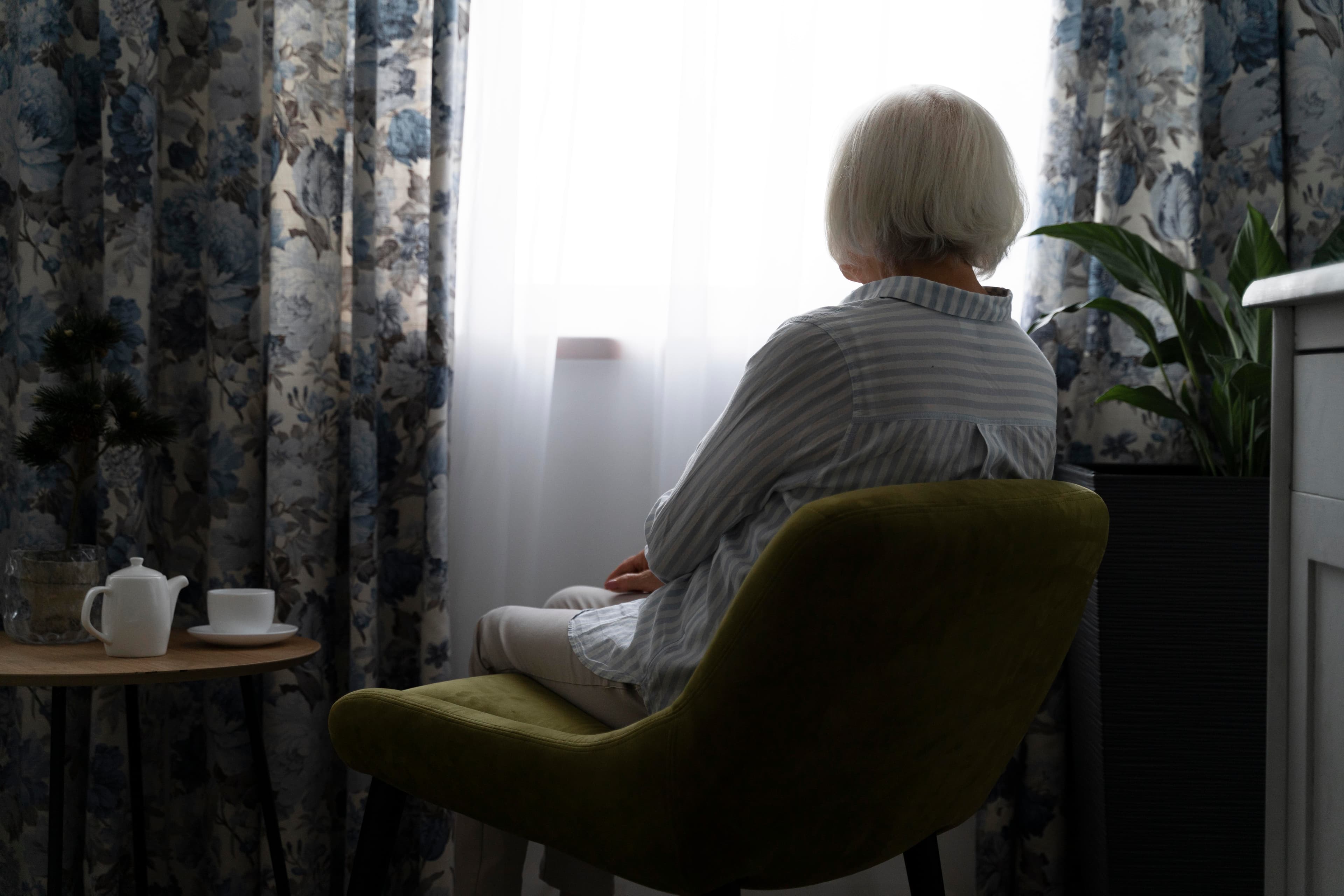 Elderly person sitting on a chair, gazing out a window with floral curtains, next to a table with a teapot and cup. - Home Instead