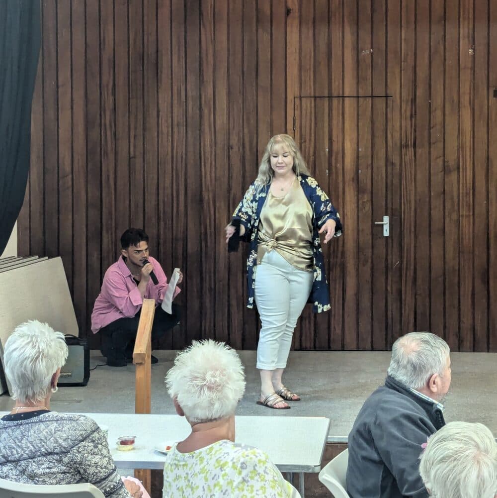 A woman in a floral jacket speaks on stage as a man observes and the audience watches in a wooden-paneled room. - Home Instead