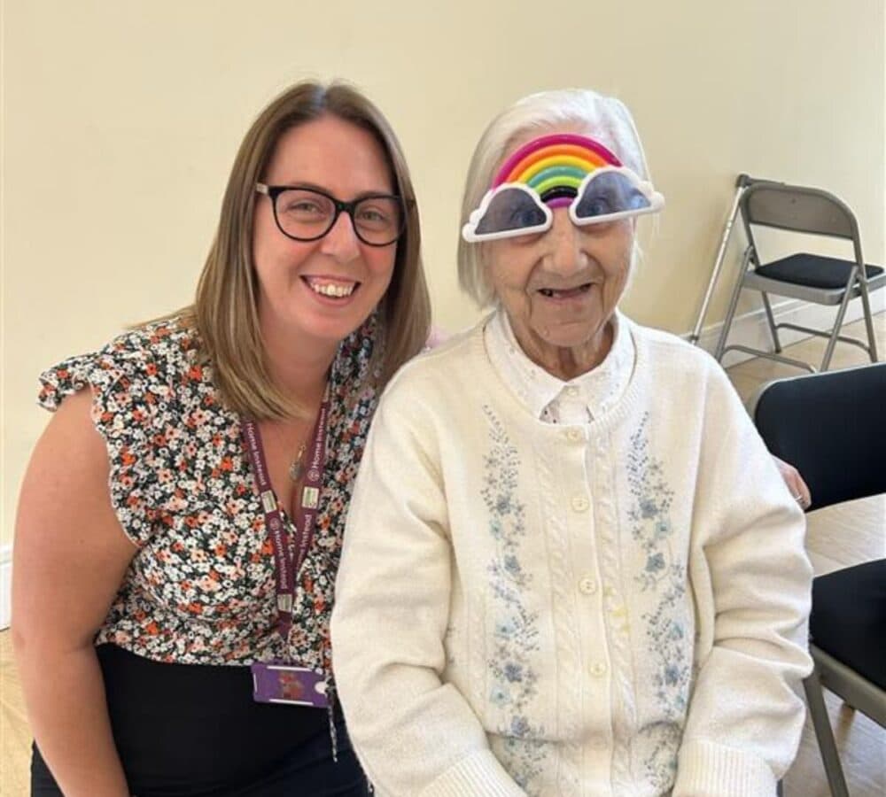 Elderly woman wearing rainbow glasses smiles with a younger woman in a floral top beside her indoors. - Home Instead