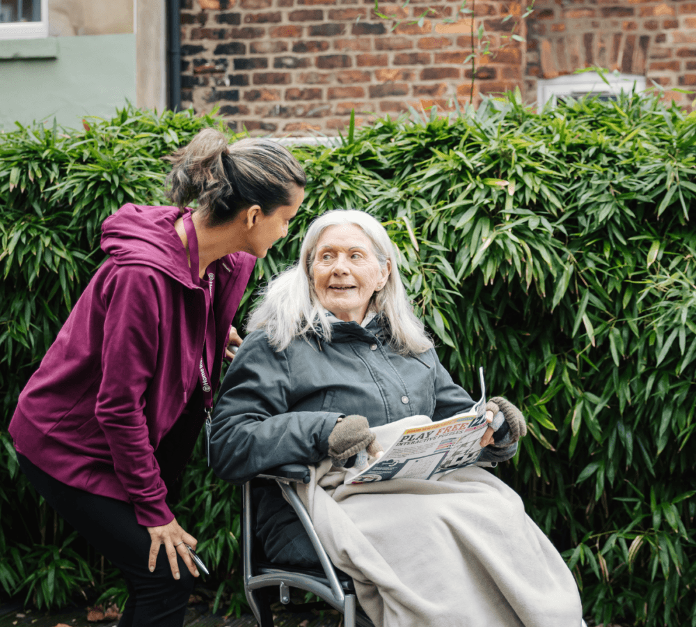 A Senior sitting on a wheelchair holding a puzzle with a Care Professional talking to her and helping her solve the puzzle
