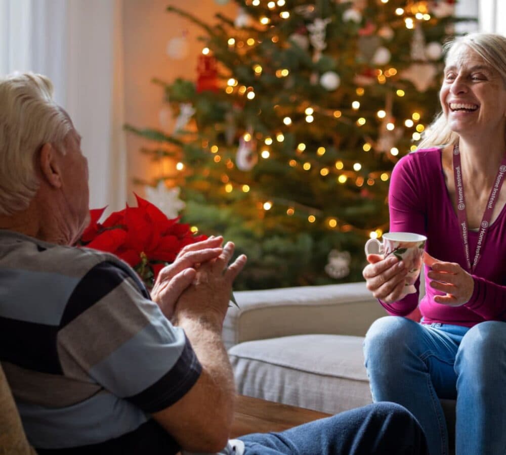 An old man with white hair sitting on a chair with a younger carer laughing while holding a cup sitting on a couch and with a Christmas tree and lights on the background