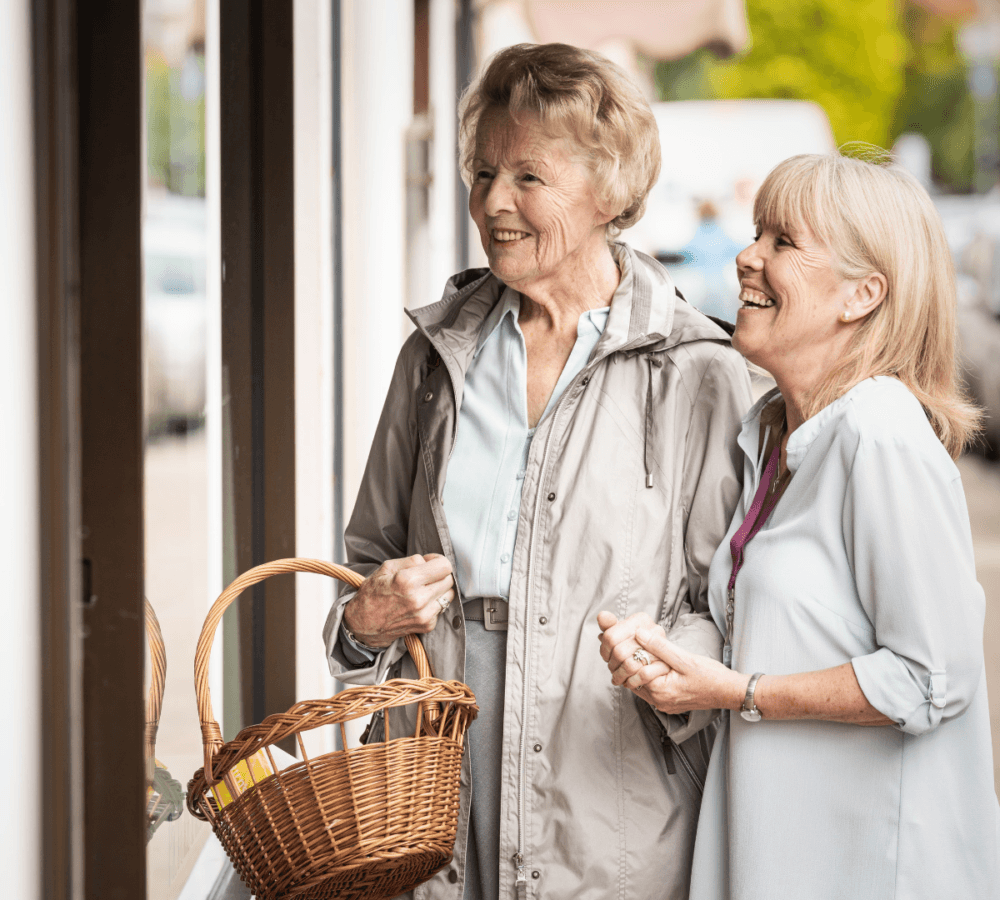 A senior woman wearing a jacket smiling and carrying a basket with another woman carer with blonde hair wearing long sleeves smiling while looking at a store window