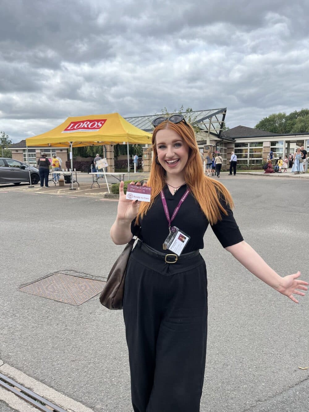 Woman smiling and holding a pass, standing in front of a yellow tent labeled "LOROS" at an outdoor event. - Home Instead