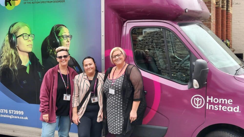 Three women smiling and posing in front of a purple Home Instead van with an autism awareness poster. - Home Instead