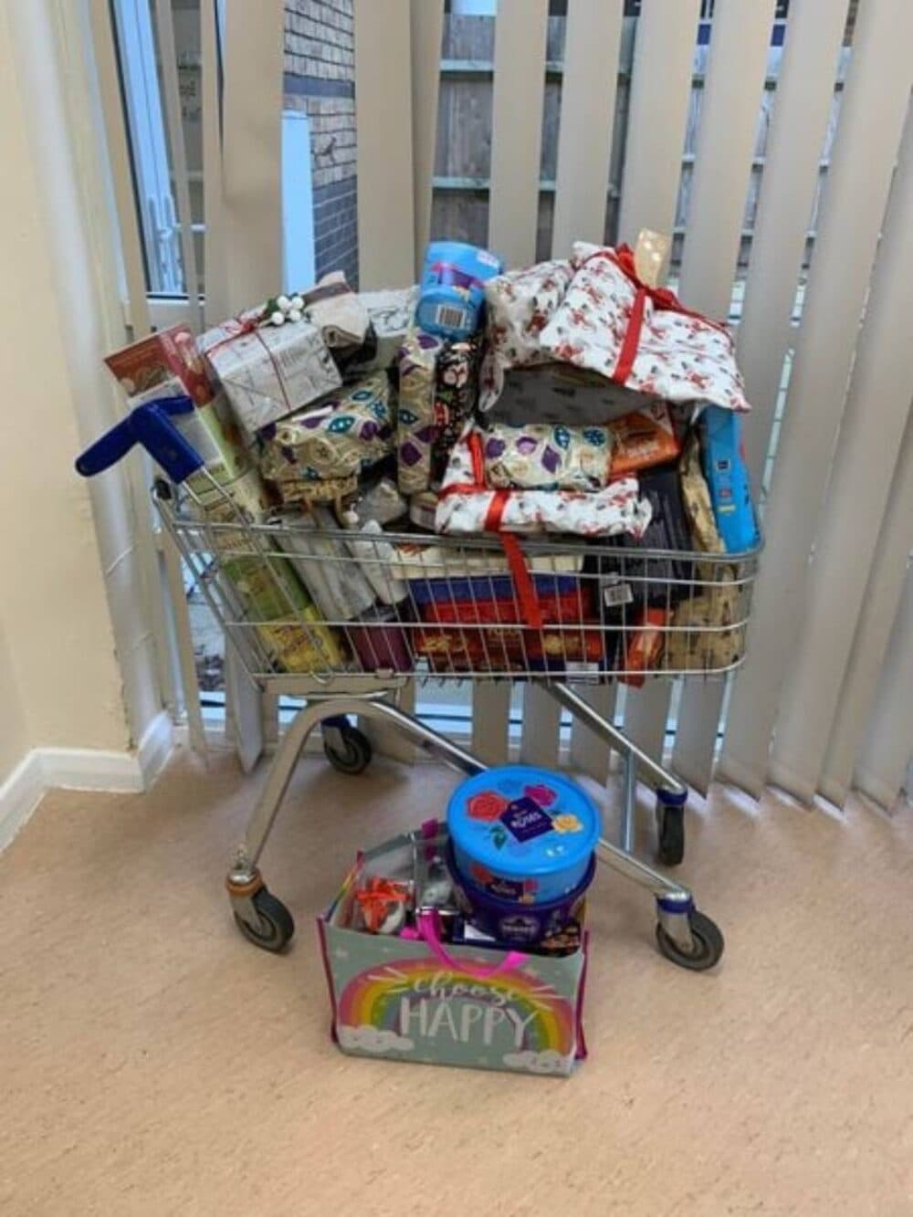 A shopping cart filled with wrapped gifts and treats, next to a colorful box labeled "HAPPY. - Home Instead