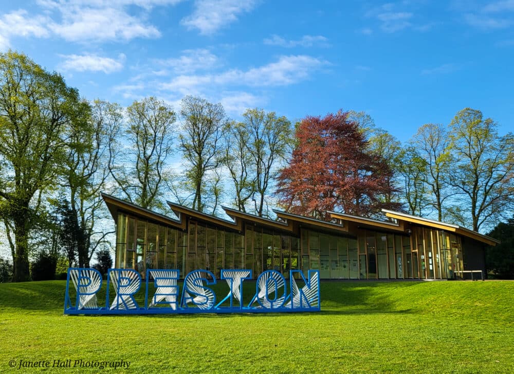 Modern building with zigzag roof behind large "PRESTON" letters on grass, surrounded by trees under a blue sky. - Home Instead