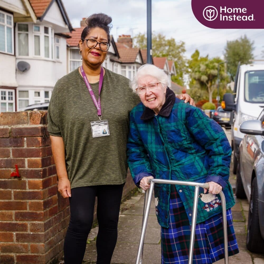 Caregiver and elderly woman with walker smiling on residential street, wearing casual clothing. - Home Instead