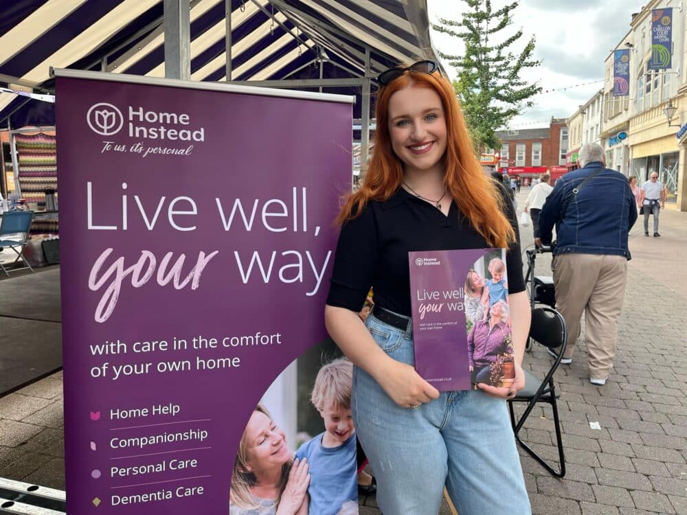 A woman holding a brochure stands by a "Home Instead" banner on a street with outdoor stalls. - Home Instead