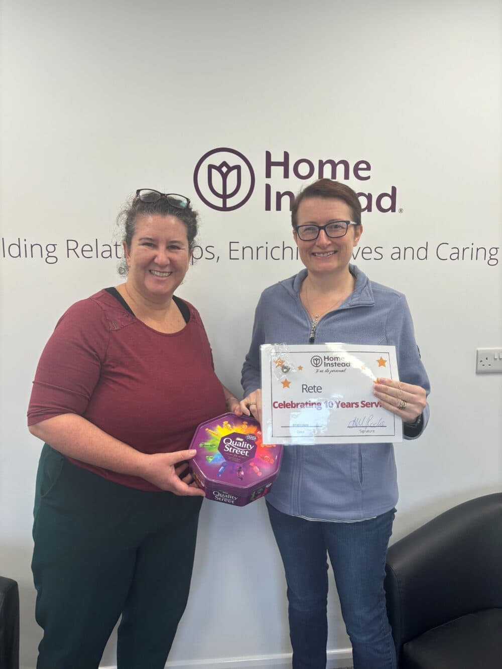 Smiling care professional holding a certificate marking her 10-year anniversary and a box of chocolates. She is standing next to Ann-Marie, Home Instead York's Managing Director, also smiling, in front of a 'Home Instead' sign.
