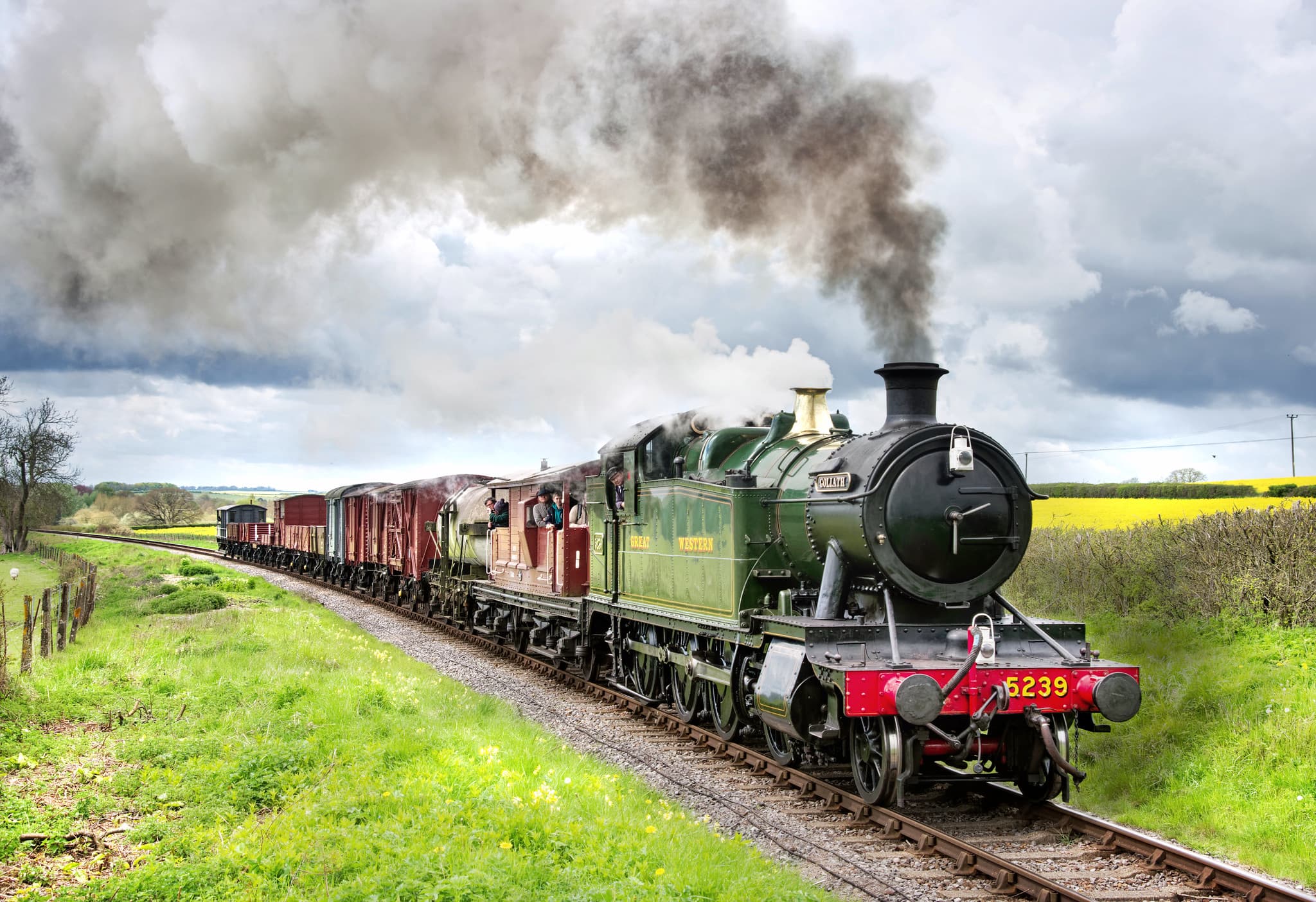 Photo of a steam passenger train moving along a scenic railway track, surrounded by the lush green countryside of Alresford. The train is green and with plumes of smoke set against a backdrop of blue sky and rolling hills.
