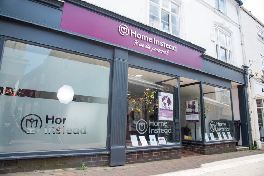 Storefront of Home Instead with logo and promotional signs on display in the windows. - Home Instead