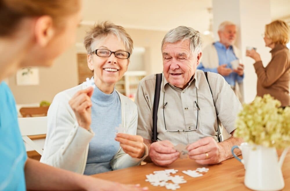 Smiling seniors assemble a puzzle at a table, with others chatting in the background. - Home Instead
