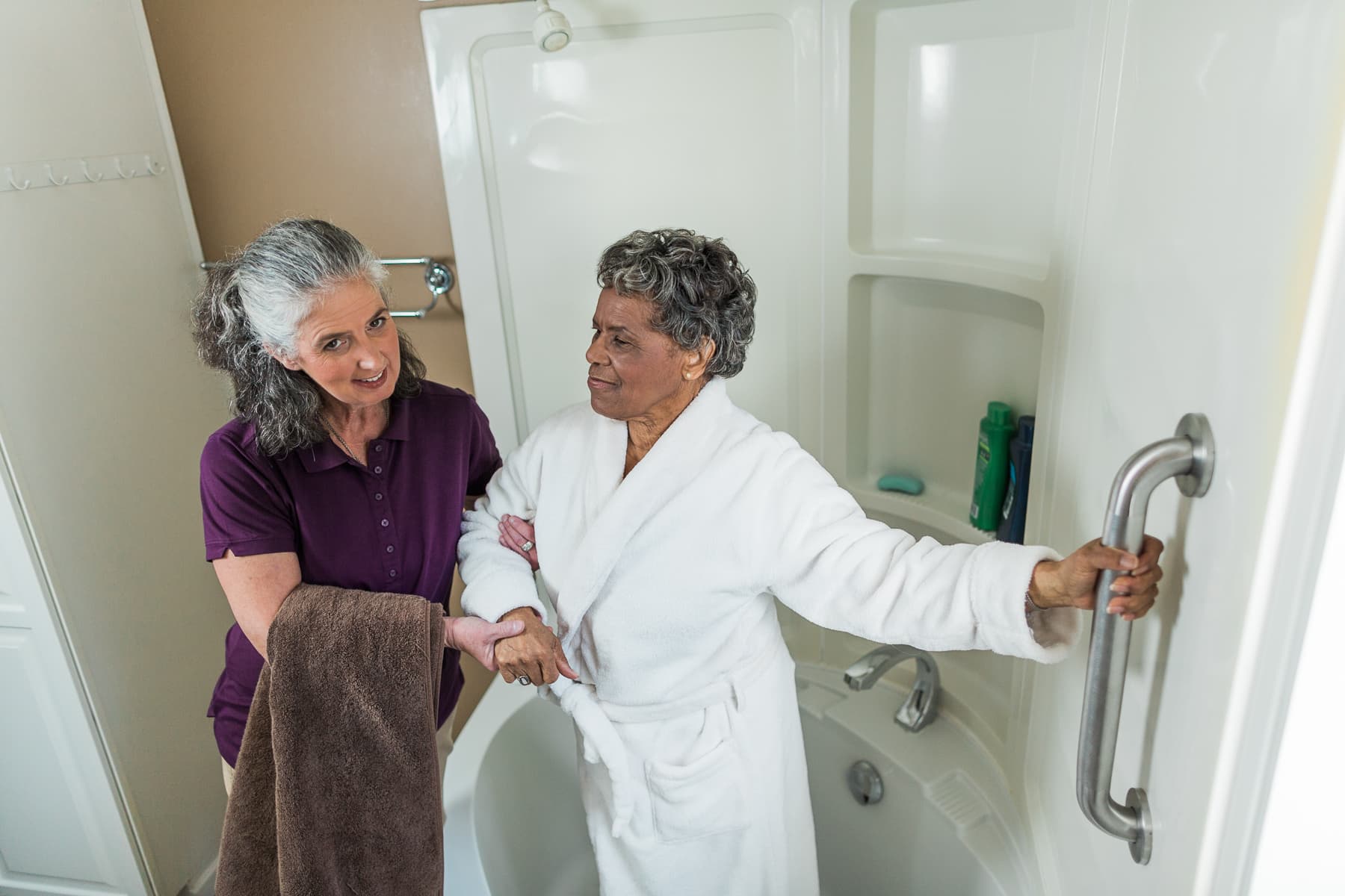 An elderly woman holds a grab bar in a bathroom while a caregiver assists her out of the tub. - Home Instead