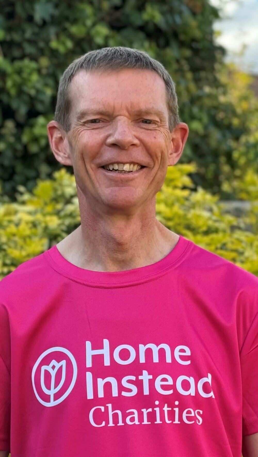 Smiling man in a pink "Home Instead Charities" shirt standing in front of green foliage. - Home Instead