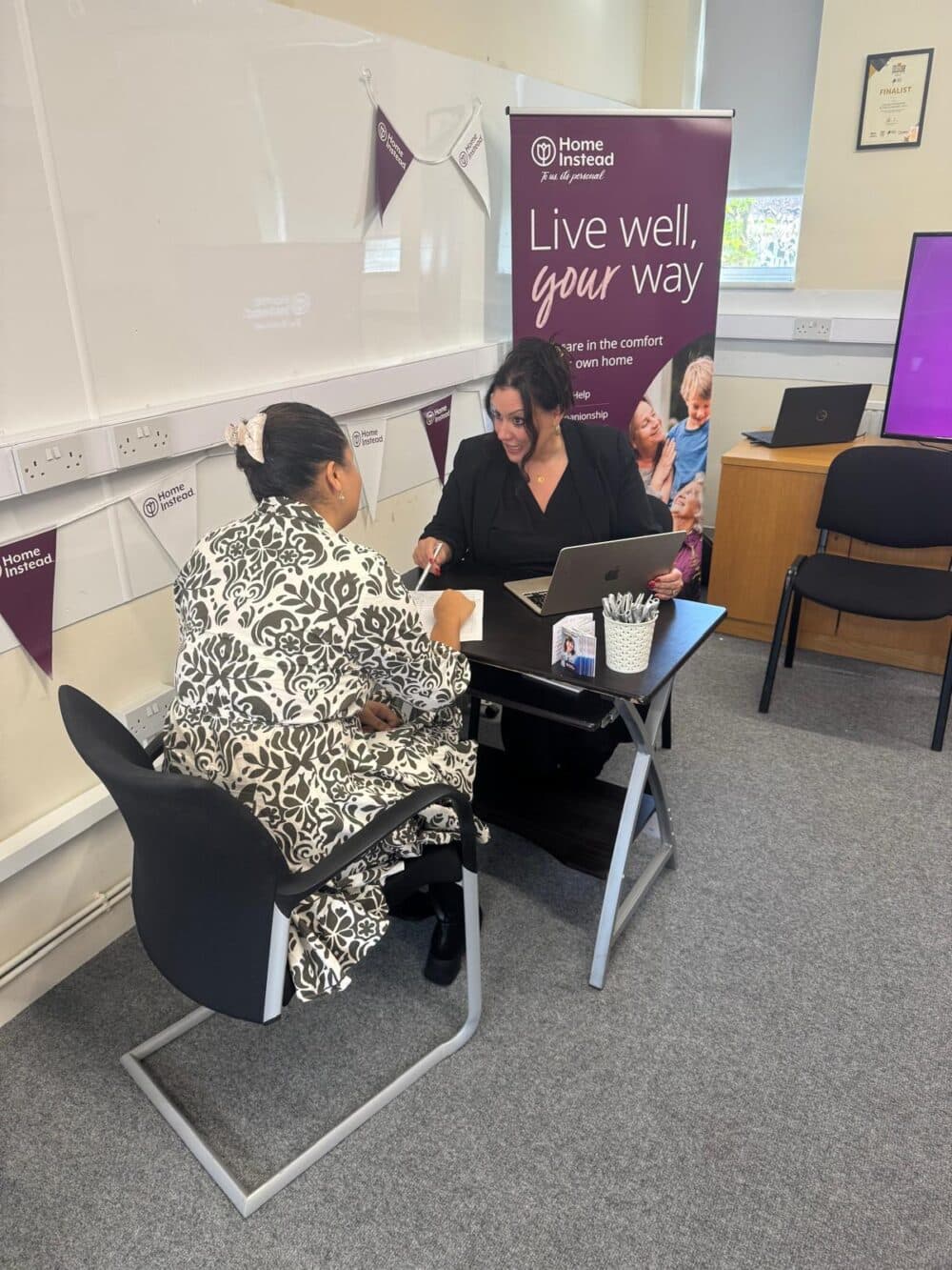 Two women sitting and talking at a desk with a "Live well, your way" banner in the background. - Home Instead