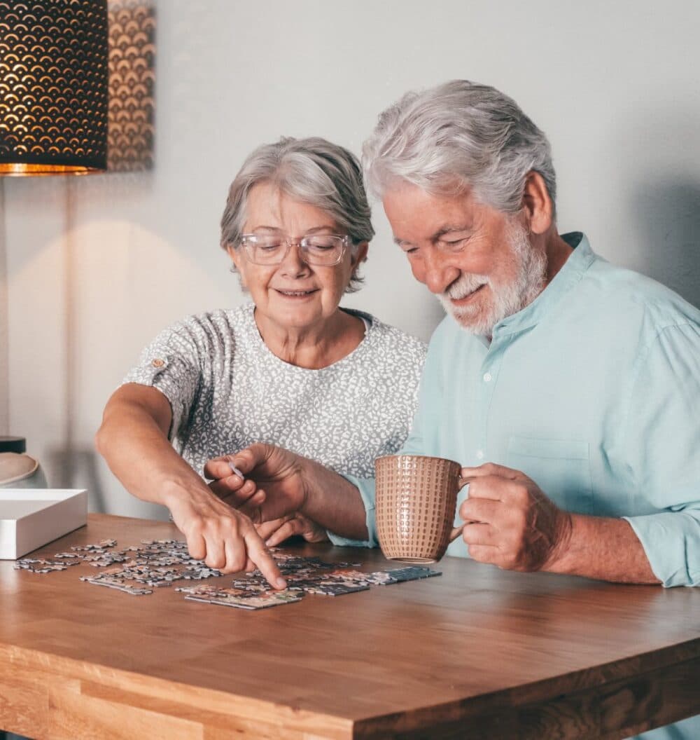 Elderly couple smiling and working on a jigsaw puzzle at a wooden table; man holds a mug. - Home Instead