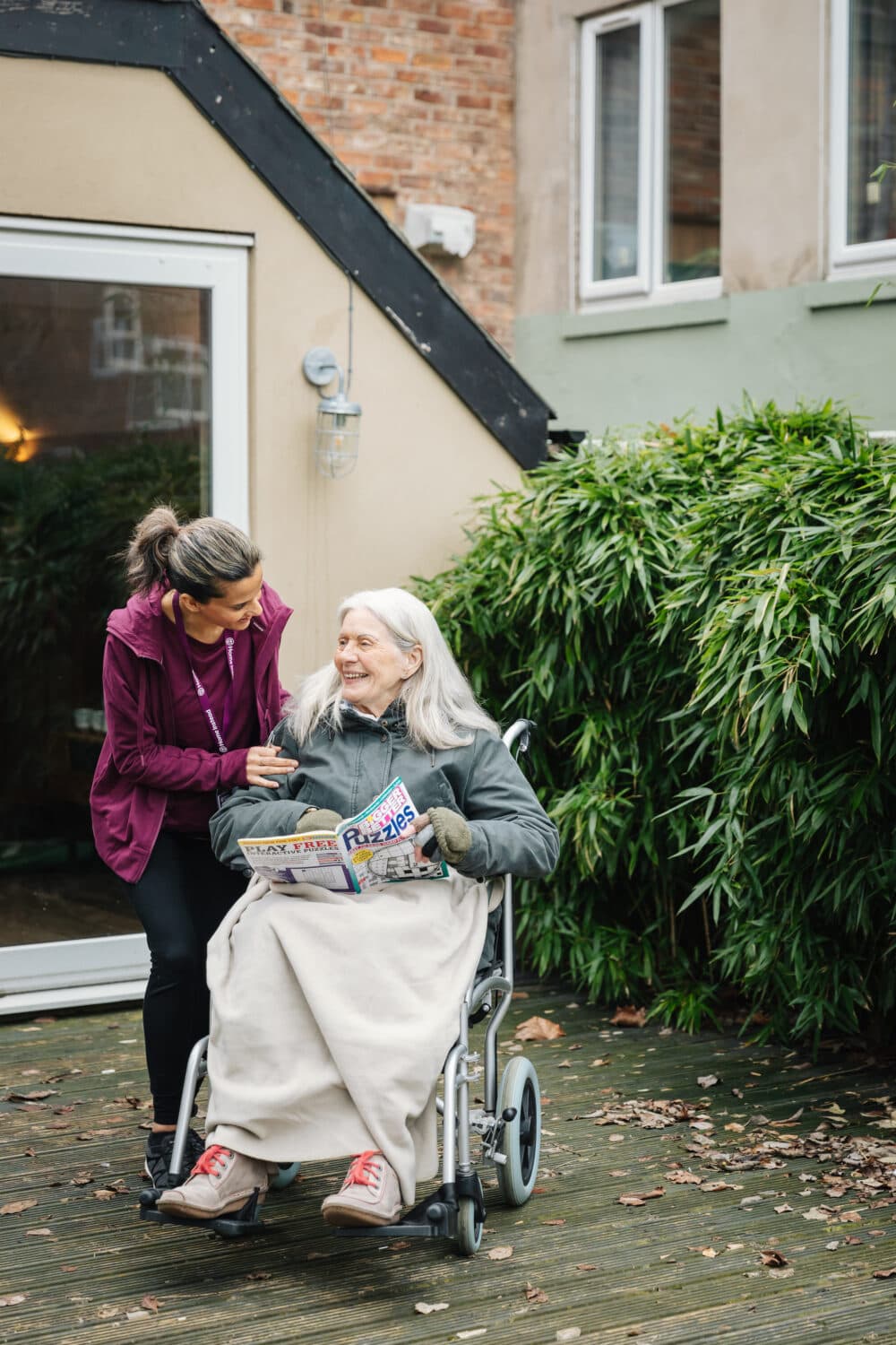 A caregiver smiles at a woman in a wheelchair reading a magazine outdoors near a house and greenery. - Home Instead
