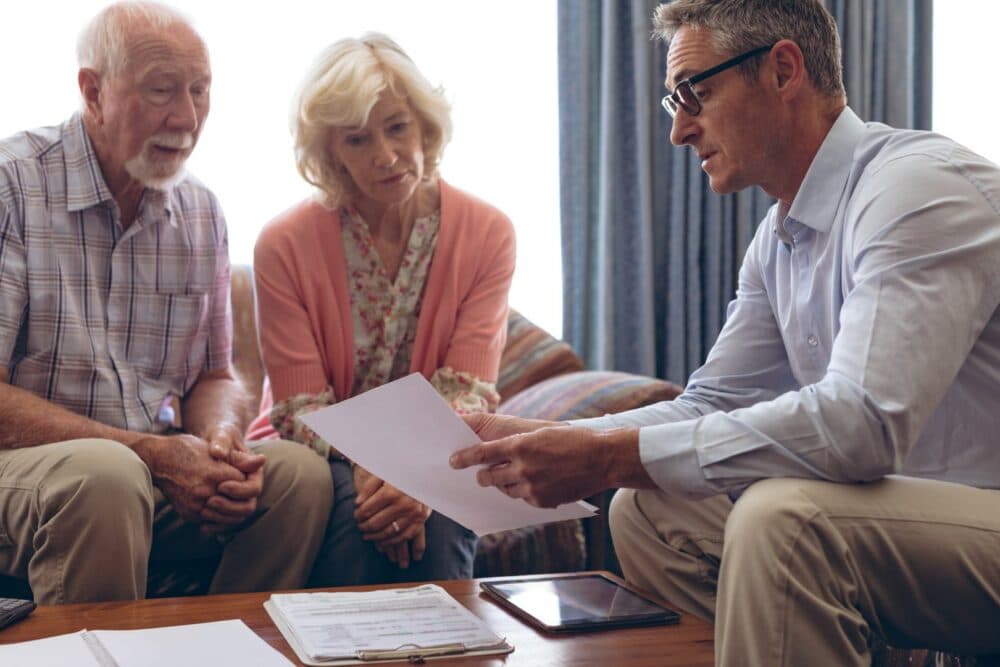A man discusses documents with an elderly couple seated on a sofa in a living room. - Home Instead