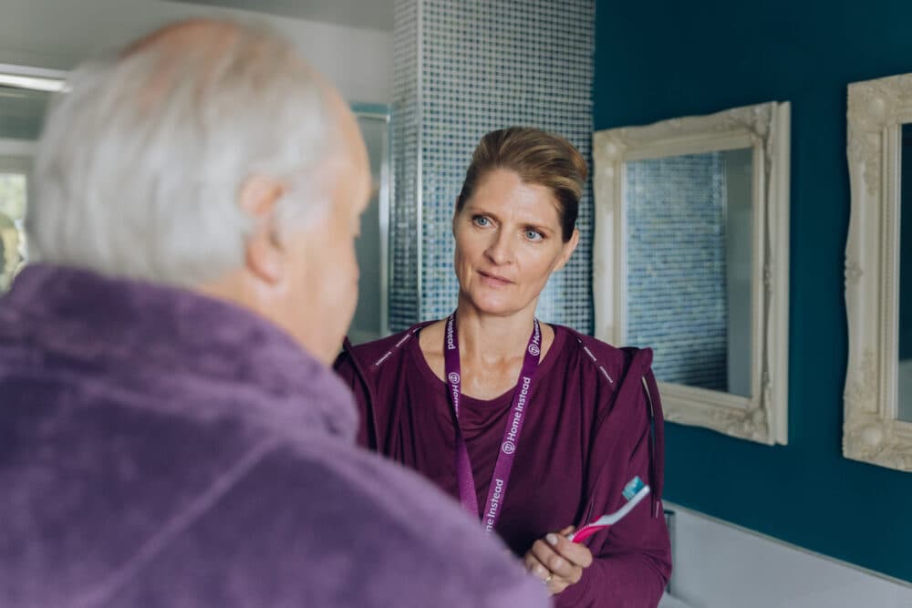 A woman wearing a lanyard talks to an older man in a bathroom, holding a toothbrush. - Home Instead