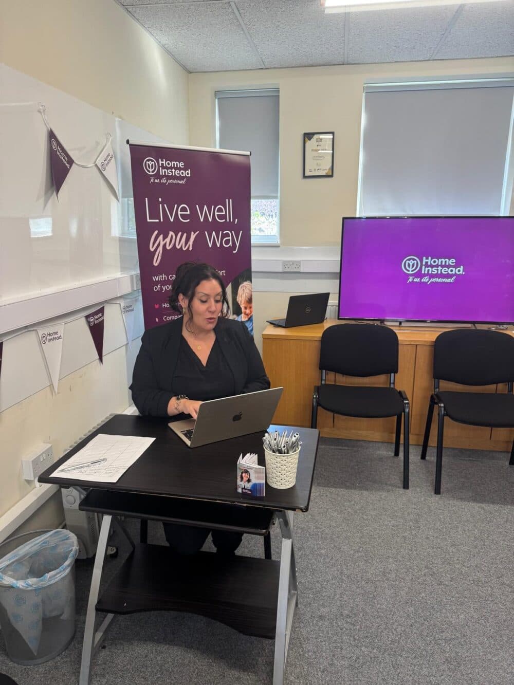 A person works on a laptop at a desk with a "Live well, your way" banner and chairs in the background. - Home Instead