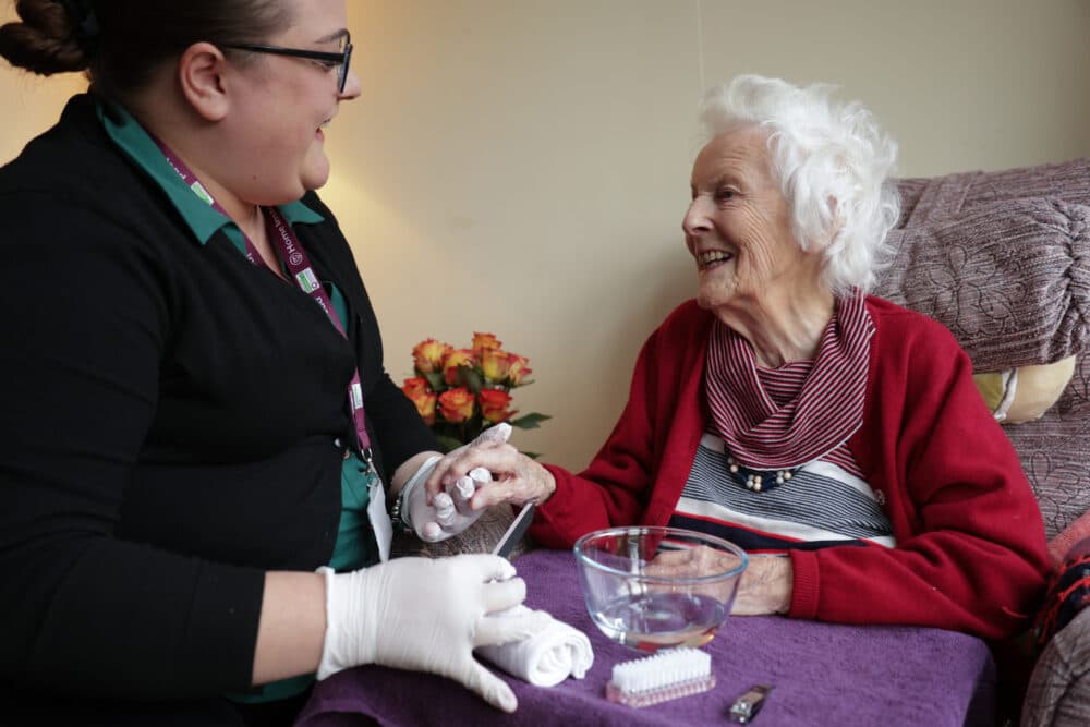A caregiver smiles while holding an elderly woman's hand during a manicure at home. - Home Instead