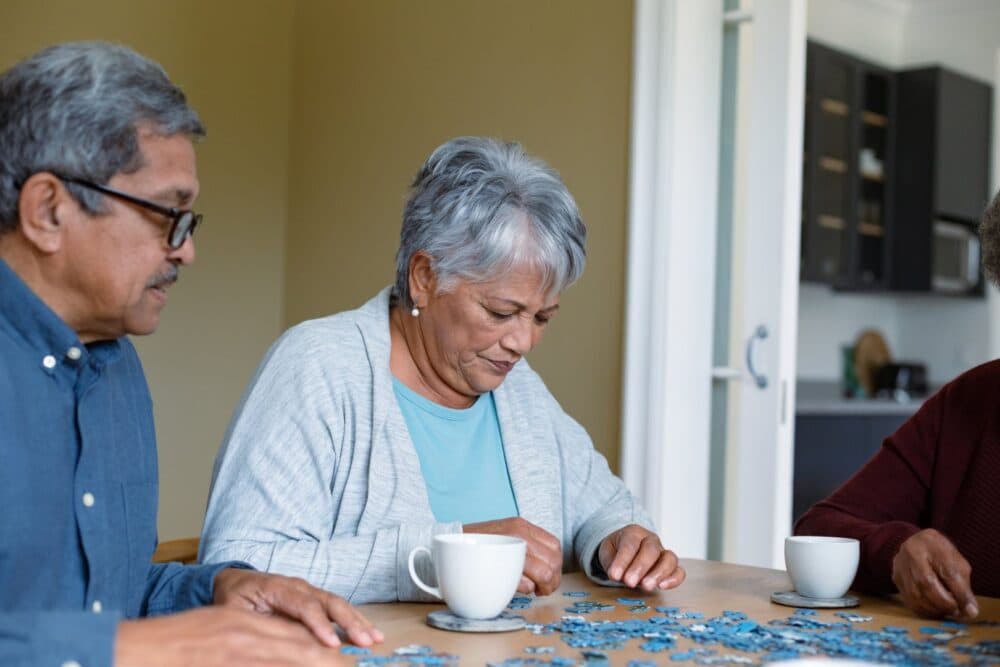 Three elderly people sit at a table working on a jigsaw puzzle with cups of coffee nearby. - Home Instead