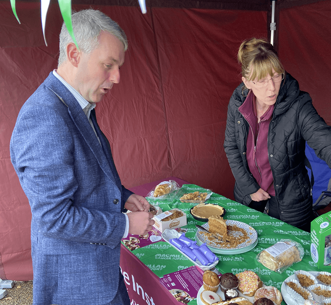 Male MP standing at Cake stand being served by lady also stood up explaining the range of cakes on sale for a donation to Macmillan.