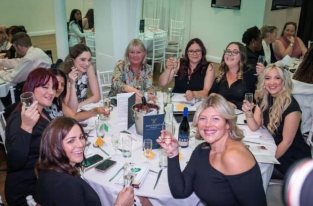 Nine women sit around a table at a formal event, smiling and raising glasses in a toast. - Home Instead