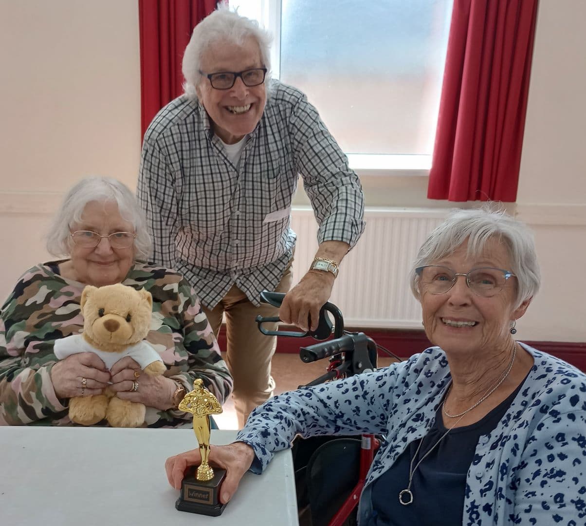 Three smiling elderly women; one holds a trophy, another a teddy bear. They gather at a table in a room with red curtains. - Home Instead