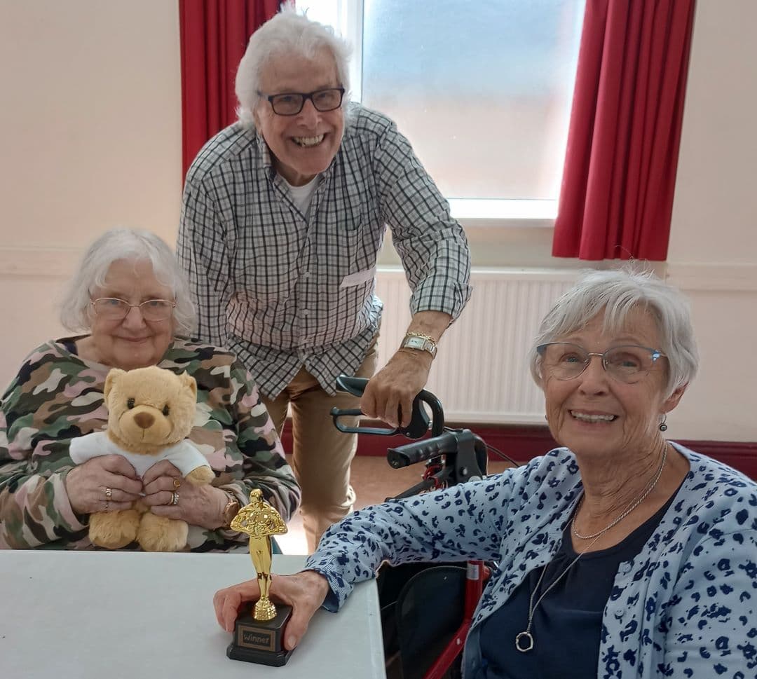 Three elderly people smile; one holds a trophy, another holds a teddy bear. They're at a table with red curtains behind. - Home Instead