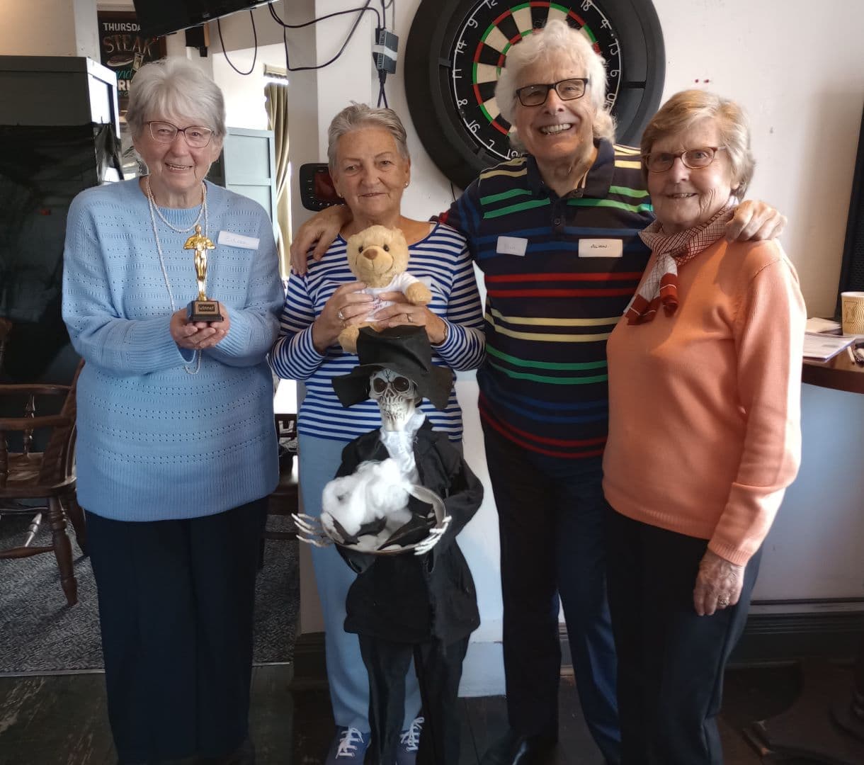 Four seniors smiling, holding a trophy and plush toys, in front of a dartboard and a skeletal decoration. - Home Instead