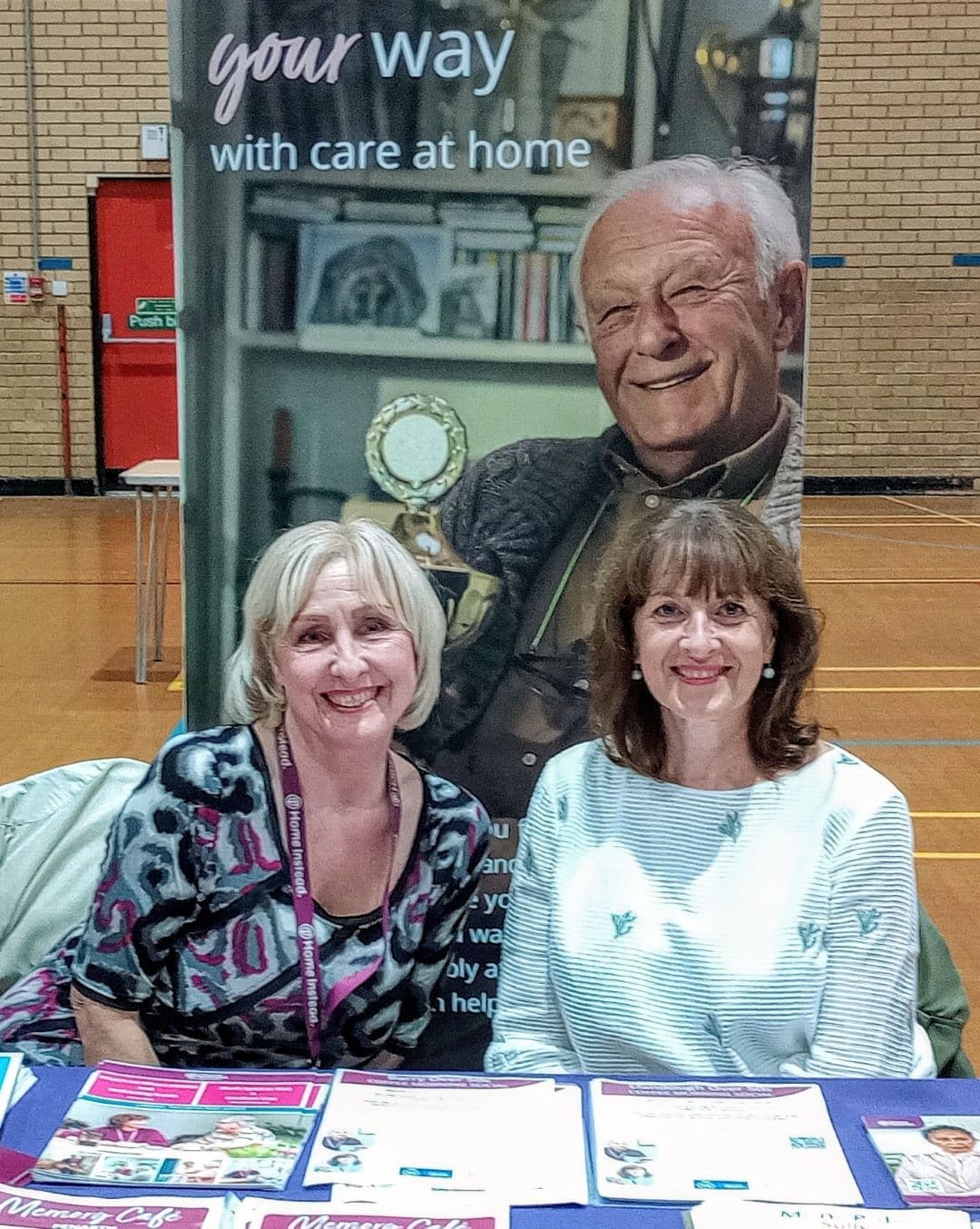 Two women at a table with brochures, smiling at an event. A poster with an elderly man is in the background. - Home Instead