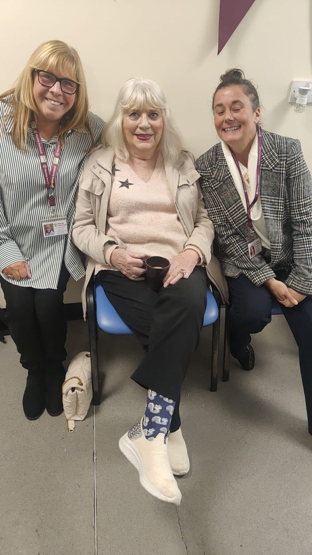 Three women sitting together; the middle one is holding a mug and wears star-patterned socks. All are smiling. - Home Instead