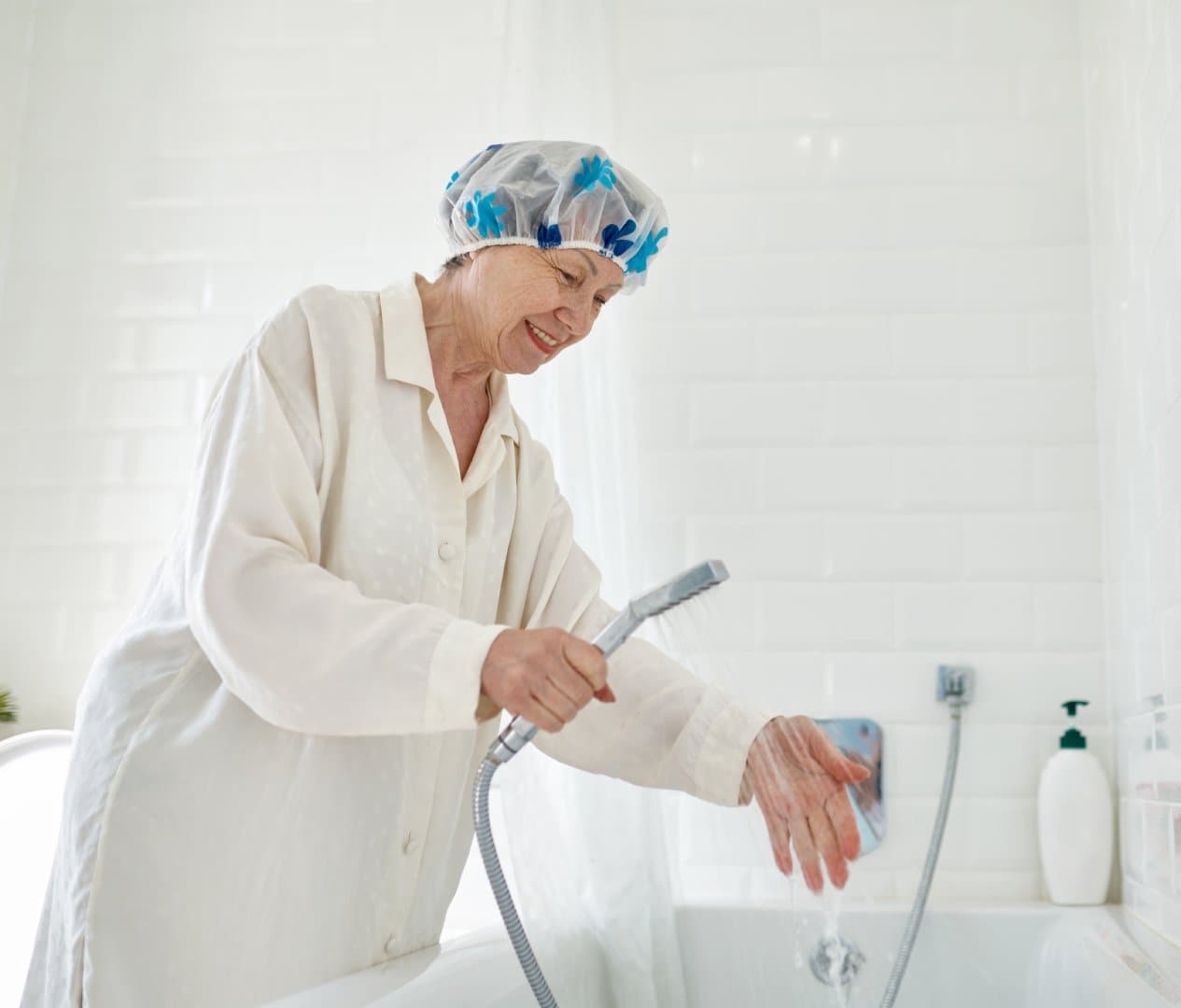 Elderly person in a shower cap and bathrobe tests water temperature with a handheld showerhead. - Home Instead
