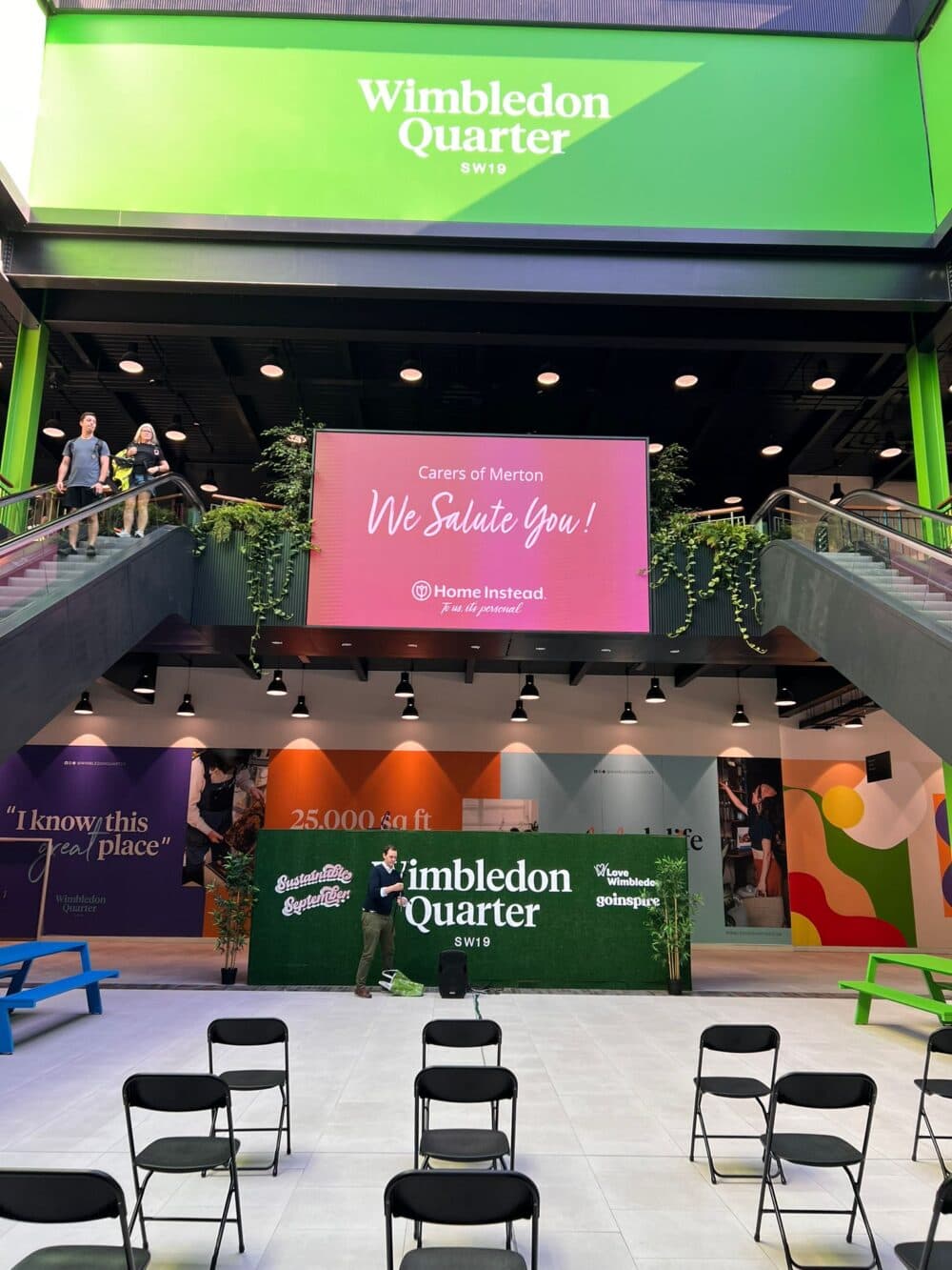 Indoor event space with a "Wimbledon Quarter" sign, a stage, and empty chairs; colorful wall art and large screen above. - Home Instead