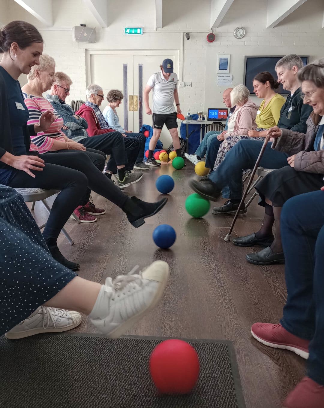 Group of people sitting in chairs in a room, passing colorful balls with their feet in a line. - Home Instead