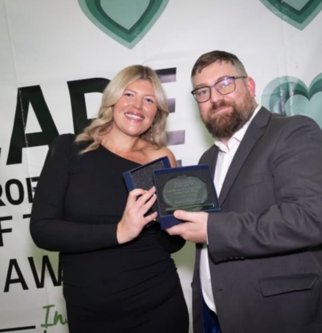 Two people smiling while holding a plaque together at an awards event with a decorated backdrop. - Home Instead