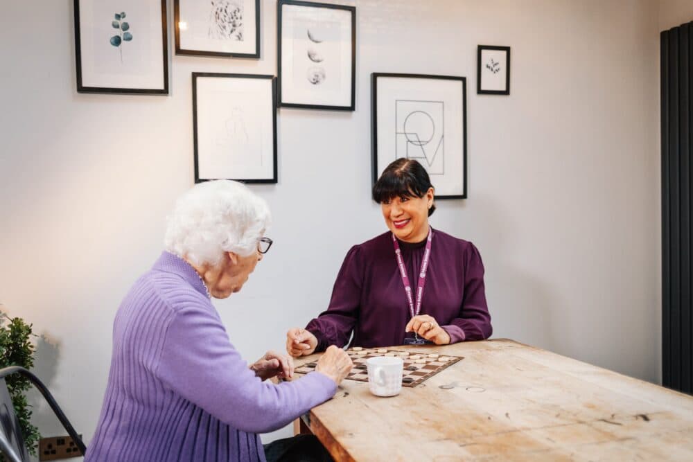 An elderly woman and a caregiver play a board game at a wooden table in a bright room. - Home Instead