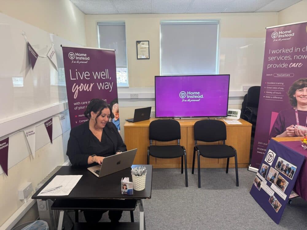 A woman working on a laptop at a desk, with Home Instead banners and a display in the background. - Home Instead