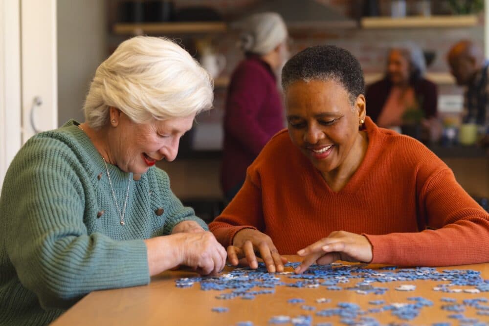 Two elderly women smiling and assembling a puzzle together at a table, with others socializing in the background. - Home Instead