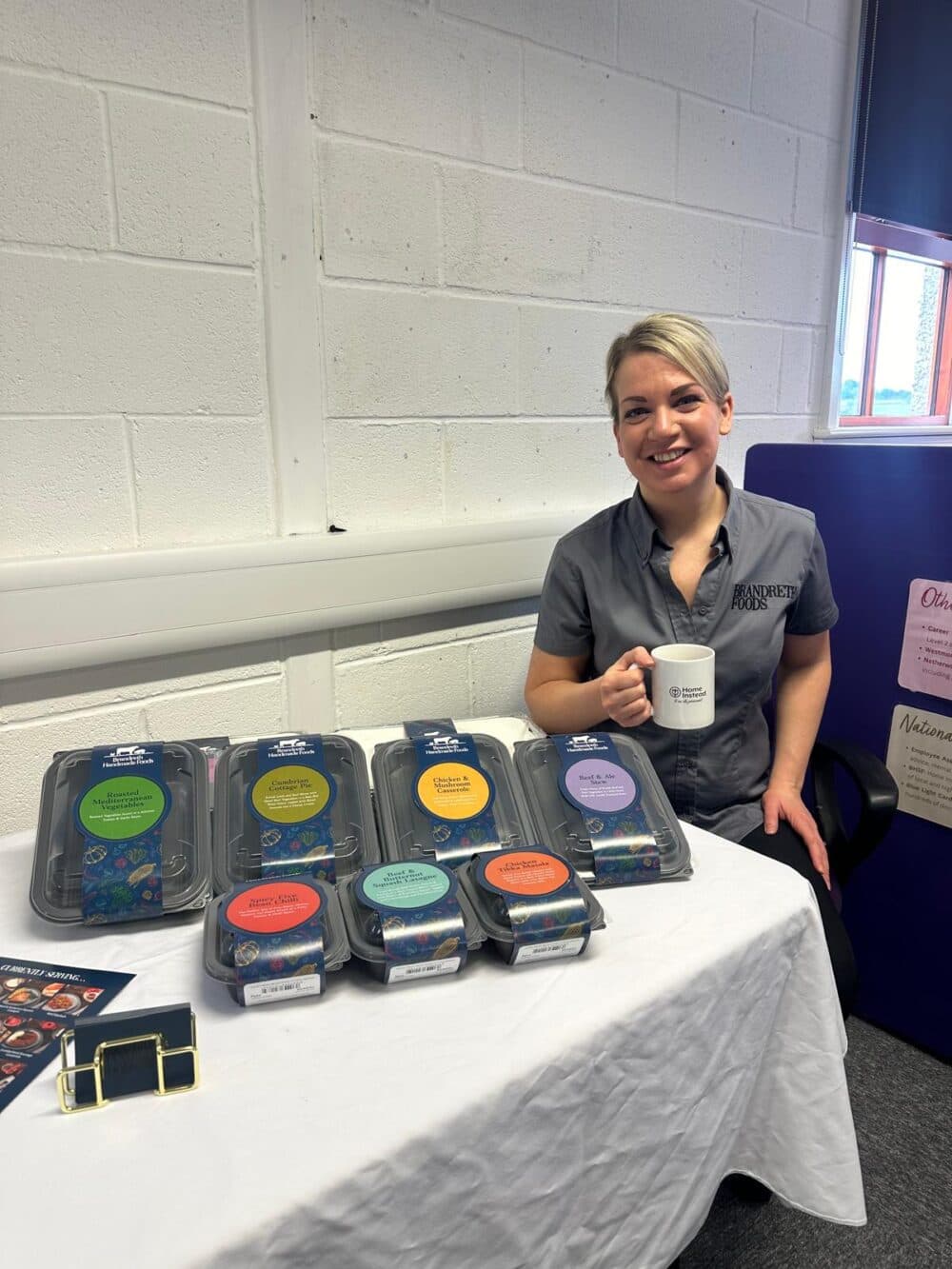 A person sits at a table with packaged products and holds a mug, smiling. - Home Instead