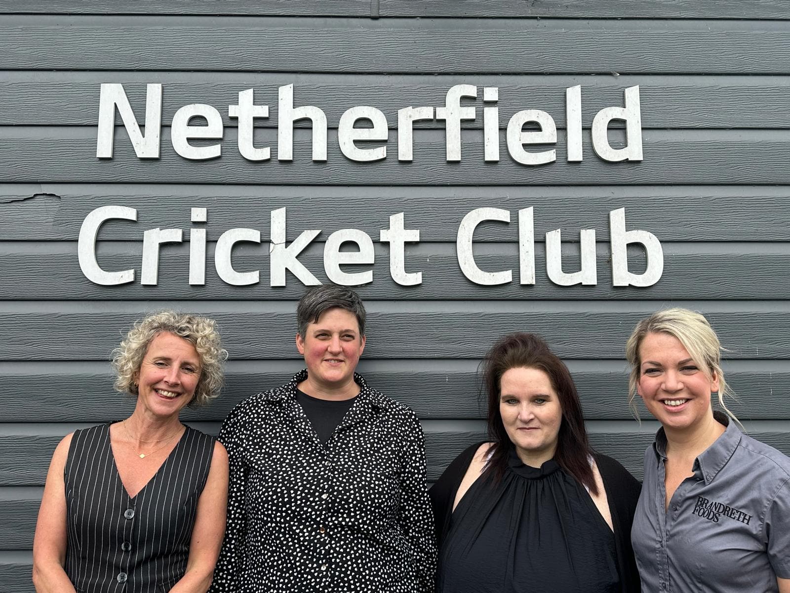 Four people smile in front of a wall with "Netherfield Cricket Club" sign. - Home Instead