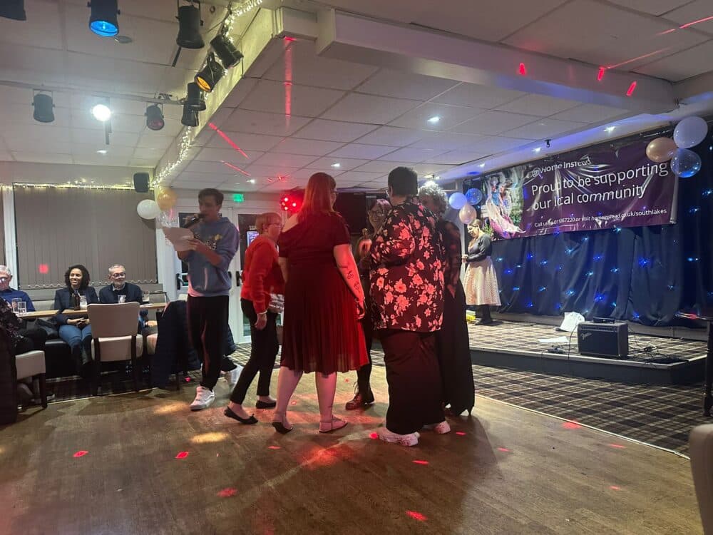 A group of people dancing on a stage at an event with a banner in the background. - Home Instead