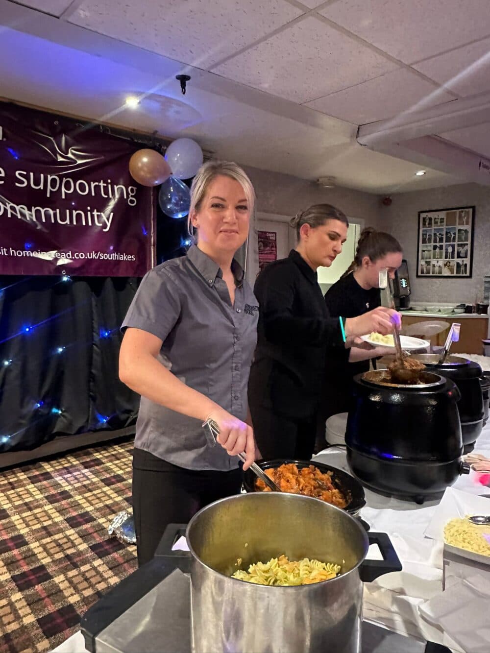 Three people serving food at an event with a banner in the background. - Home Instead