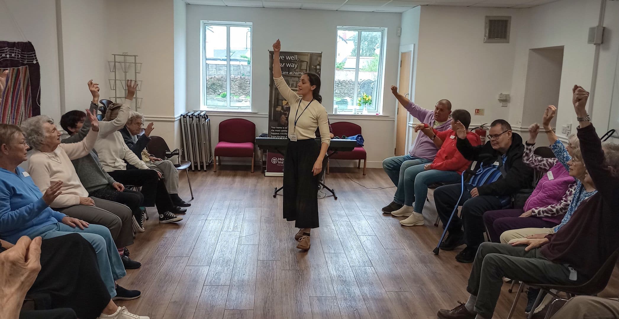 A woman leads a seated exercise class for seniors in a bright room, with participants raising their arms. - Home Instead