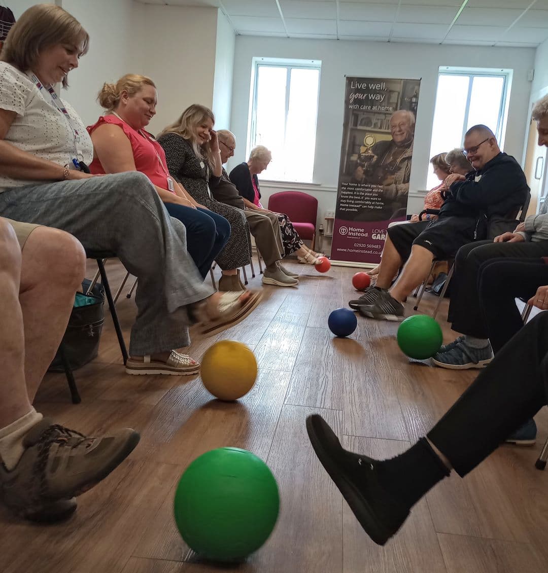 A group of people sitting in a circle, engaging in a seated exercise with colorful balls on the floor. - Home Instead