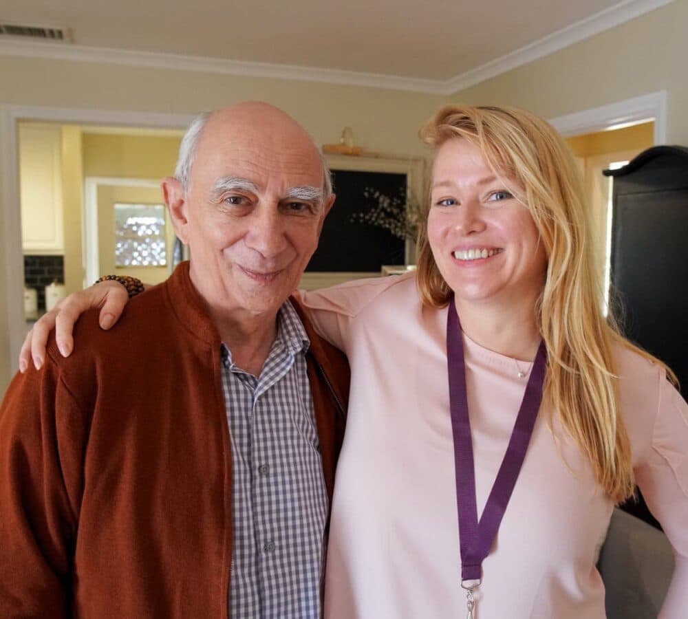 A happy male senior standing and smiling with his younger carer with long hair and smiling inside the house